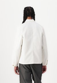 Persona con trenzas que lleva una chaqueta blanca y pantalones gris oscuro, de pie de espaldas contra un fondo blanco liso.