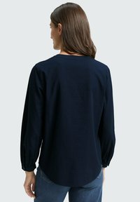 Camicia a maniche lunghe blu navy con tessuto testurizzato, scollatura rotonda e polsini arricciati, abbinata a jeans blu chiaro. Vista posteriore mostrata.