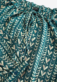 Teal-mönstrad blus i slät tyg, med rynkad halsringning, snörning och dekorativa knappar med bladmönster.