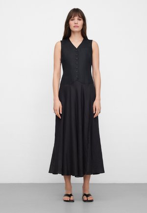 Kobieta stoi przy białej ścianie, ubrana w czarną sukienkę midi bez rękawów z guzikami oraz czarne sandały z odkrytymi palcami, skierowana przodem z neutralnym wyrazem twarzy.