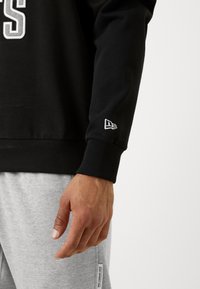 Felpa nera con polsini a costine e un logo prominente sul petto. Il polsino presenta un piccolo logo ricamato. Pantaloni della tuta grigi parzialmente visibili.