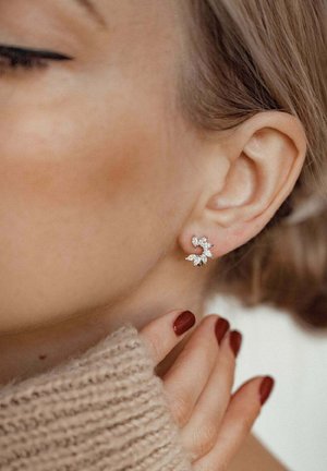 Pendientes plateados en forma de flor adornados con piedras preciosas transparentes, mostrando intrincados diseños de pétalos. Una mano toca suavemente el pendiente.