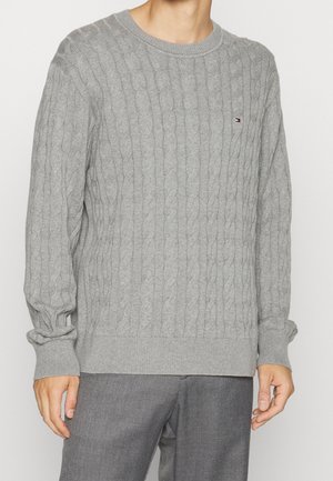 Mann trägt einen hellgrauen Pullover mit Zopfmuster, gerippten Bündchen und Saum, kombiniert mit grauen Hosen vor einfachem Hintergrund.