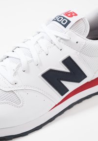 Sneaker bianche New Balance con logo "N" navy, accento rosso sulla suola intermedia, tomaia in mesh e pelle, e lacci bianchi.