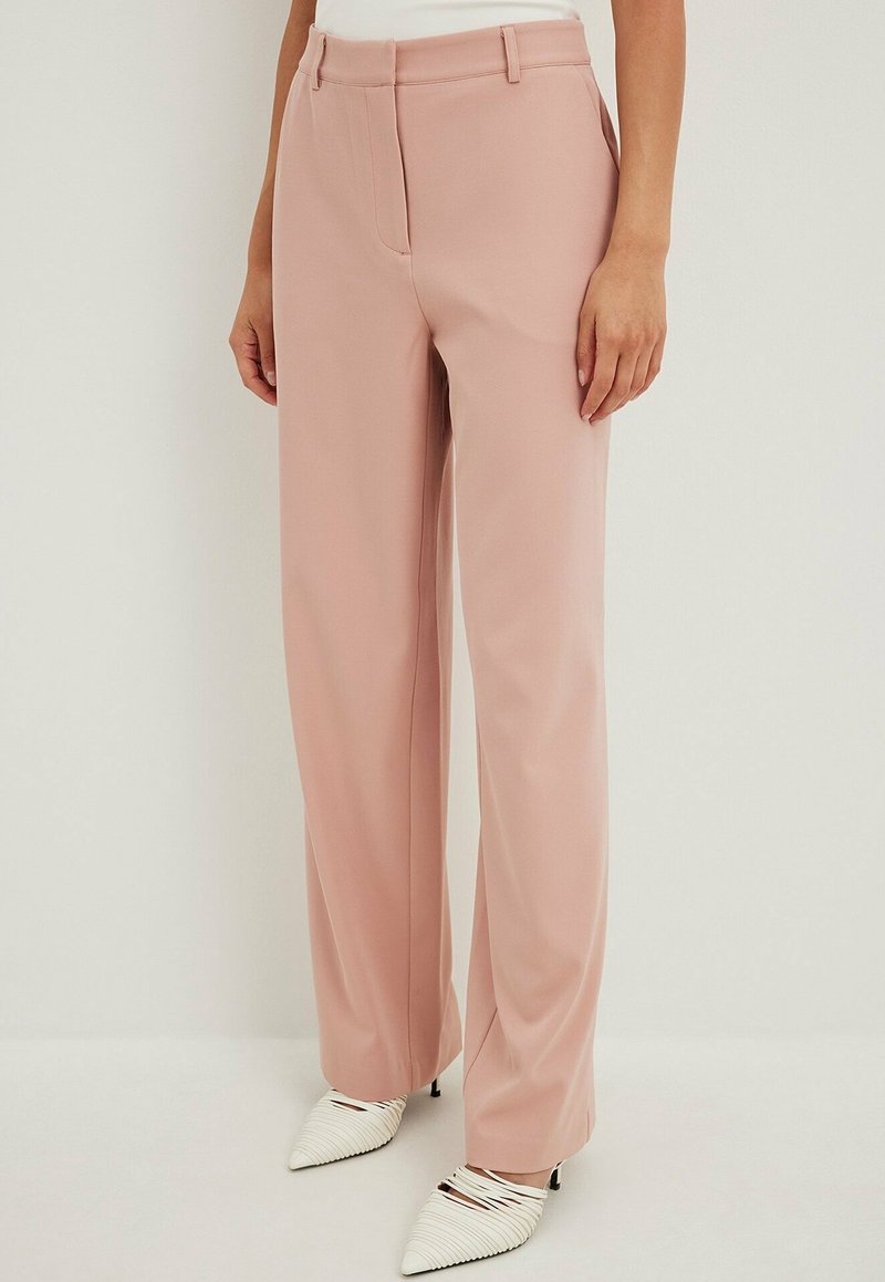 Woman wearing high-waist, wide-leg blush pink trousers paired with white pointed-toe heeled shoes against a plain background.