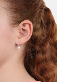 Orecchino a cerchio in argento con un piccolo charm a forma di stella, esposto su uno sfondo di capelli ricci di colore rosso-bruno e pelle.