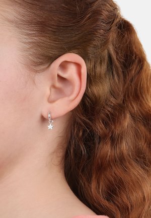 Orecchino a cerchio in argento con un piccolo charm a forma di stella, esposto su uno sfondo di capelli ricci di colore rosso-bruno e pelle.