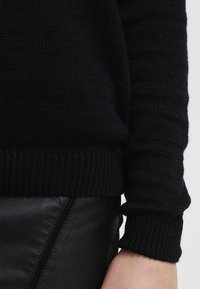 Gros plan d'une personne portant un pull en tricot noir et un pantalon en cuir noir, montrant le bas du torse et le bras.