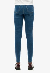 Jeans ajustados azules de mezclilla, con una textura suave, dos bolsillos traseros y un corte ceñido hasta los tobillos. Combinados con zapatos negros.