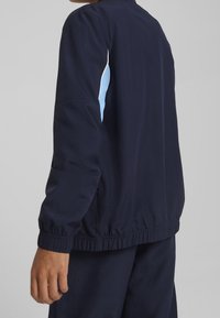 Veste légère bleu marine avec des poignets et un ourlet élastiques, présentant un accent bleu clair le long de l'épaule. Texture lisse et mate.