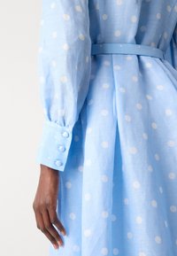 Light blue dress with white polka dots; features long sleeves with buttoned cuffs and a tied waist belt. Soft, woven fabric texture.