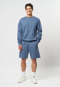 Lichtblauwe sweatshirt en bijpassende shorts gemaakt van een zacht materiaal. Voorzien van ribbels aan de mouwen en een geborduurd logo op de shorts.