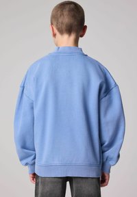 Sudadera de gran tamaño en azul claro con cuello y puños acanalados; textura suave; la vista posterior muestra un acabado liso con hombros caídos.