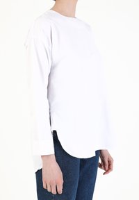 Hvid langærmet skjorte med rund halsudskæring, sideslidser og en let hi-low kantdesign, parret med mørkeblå jeans.