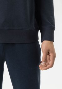 Marineblauwe sweatshirt en broek van zachte stof; close-up van een hand die tegen de dij rust; geribbelde manchetten en zoomdetails zichtbaar.