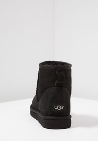 Botín de ante negro con un suave forro interior, detalles de costura en el lateral y un logo de UGG en la parte posterior. Suela de goma plana.