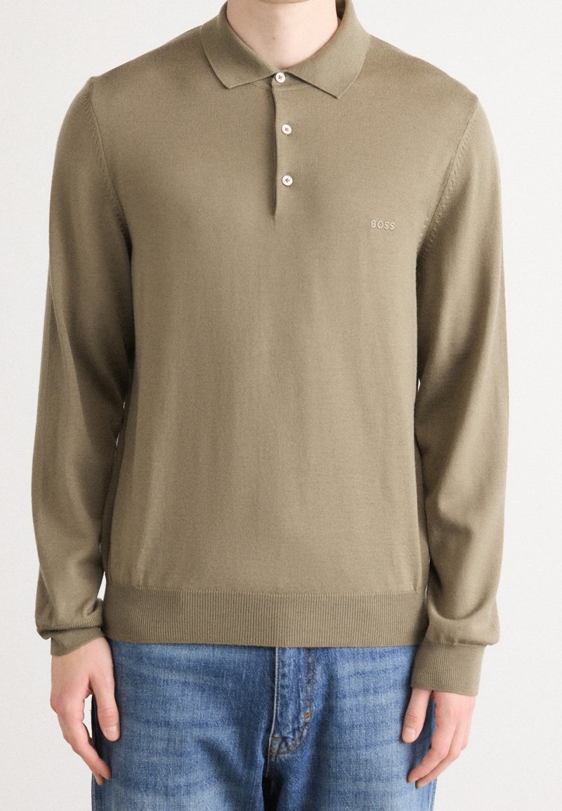 Homme portant un pull polo à manches longues vert olive avec trois boutons et un logo brodé clair, associé à un jean bleu.