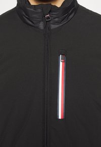 Black jacket with a high collar, zippered front, and a chest pocket featuring red, white, and blue accents on a black background.