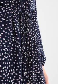 Abito floreale navy con piccoli fiori rossi e bianchi, maniche lunghe, vita regolabile con coulisse e dettagli in bottone lungo la parte frontale.