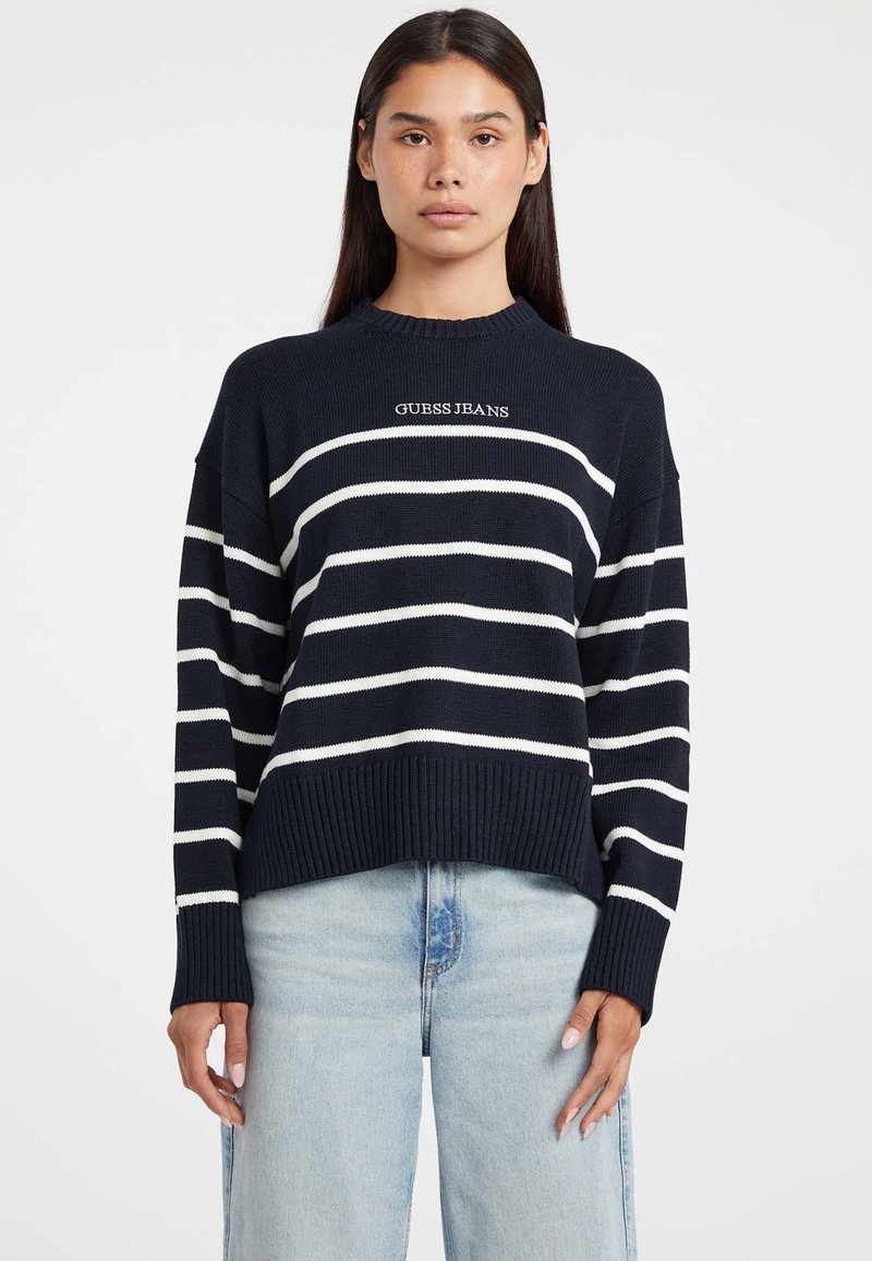 Femeie îmbrăcată cu un pulover bleu închis cu dungi orizontale albe și textul "GUESS JEANS", asortat cu blugi din denim bleu deschis.