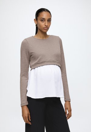 Kvinde med glat, mørkt hår iført en taupe ribstrikket langærmet sweater lag-på-lag over en hvid bluse og sorte bukser, stående foran en ensfarvet baggrund.
