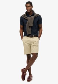 Marine polo skjorte, lys khaki shorts, brun bælte, brun sweater draperet over skuldrene, bordeaux sejlersko med snørebånd, glat tekstur.
