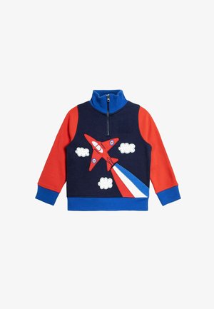 Bluza z suwakiem, z granatowym korpusem, czerwonymi rękawami i niebieskimi akcentami. Posiada haftowaną czerwoną samolot i białe chmury.