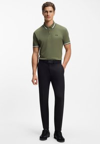 Zielona koszulka polo z białym kołnierzykiem w paski, czarne spodnie i czarne tenisówki. Na koszulce i pasku logo „BOSS”. Gładka faktura materiału.