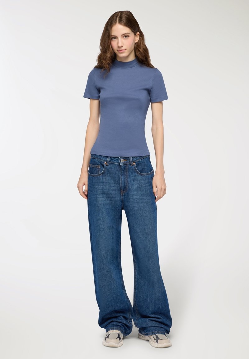 Jasnoniebieski dopasowany t-shirt z krótkim rękawem i stójką, zestawiony z luźnymi niebieskimi dżinsami oraz szarymi sneakersami. Gładka faktura, casualowy design.