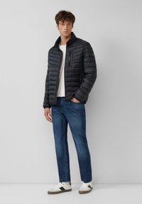 Zwarte donsjas met een opstaande kraag, een ritszak op de borst en horizontale doorgestikte naden. Gecombineerd met een blauwe spijkerbroek en witte sneakers.