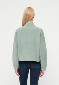 Femme aux cheveux blonds raides portant une veste en velours côtelé vert clair et un jean bleu foncé, debout de dos devant un fond uni.