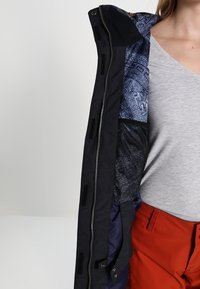 Navy buitenjack met een geometrisch patroon op de blauwe binnenvoering, ritssluiting en zakken. Soepele stof met een matte afwerking.