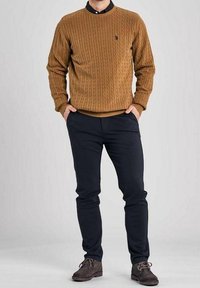 Man som bär en brun kabelstickad tröja över en svart kragskjorta, mörka slim-fit byxor och mörka mockaskor, med händerna i fickorna.