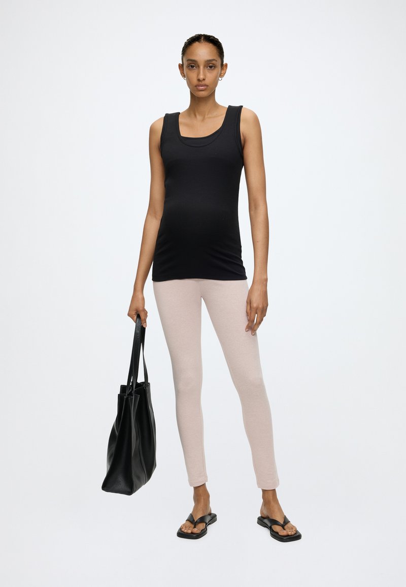 Kvinde står med ansigtet fremad, iført sort tanktop, lyse beige leggings, sorte sandaler og holder en stor sort totebag.