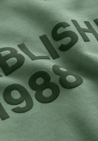 Zelena pamučna tkanina s velikim, tamno maslinastim tekstom koji kaže "OSNOVANO 1988." Materijal ima glatku teksturu s mekim završetkom.