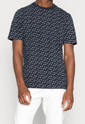 Man draagt een marineblauw T-shirt met korte mouwen, kleine witte abstracte stippen en een witte broek tegen een effen achtergrond.