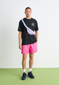 Mand står på grøn gulv iført sort t-shirt, lyserøde shorts, sorte sneakers med blå såler, hvide sokker og en lavendel crossbody taske.