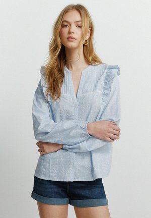 Młoda kobieta o długich blond włosach, ubrana w jasnoniebieską bluzkę z falbankami i ciemne krótkie spodenki z denimu, stojąca z założonymi ramionami.