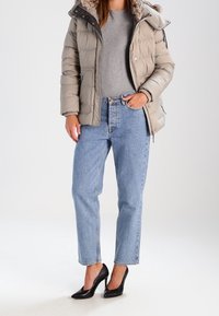 Daunenjacke in Hellgrau mit gefütterter Kapuze aus Fell, graues eng anliegendes Shirt, hochtaillierte blaue Jeans und schwarze High-Heels.