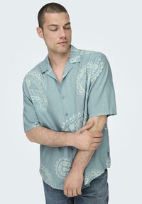 Kurzärmliges blaues Hemd mit einem hellen Paisleymuster, Knopfleiste und weichem Material. Getragen über verwaschenen Jeans mit einem lässigen Schnitt.