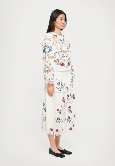 Valkoinen kukkakirjailtu mekko pitkähihaisilla hihoilla, pitsikoristeilla ja kerroksellisella hameella, yhdistetty mustiin ballerinoihin. Elegantti muotoilu eloisilla kuvioilla.