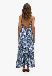 Robe maxi bleue et blanche, avec un motif floral, des bretelles spaghetti réglables, un dos nu avec des nœuds et un ourlet à volants.