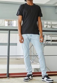 Camiseta negra de manga corta con hombros texturizados, jeans azul claro y zapatillas en blanco y negro. Ajuste casual con detalles de diseño mínimos.