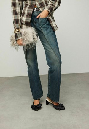 Personne portant un jean bicolore bleu, une chemise à carreaux marron, des chaussures à talons noires avec des nœuds, tenant un sac à main gris duveteux, debout sur un sol gris.