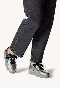 Baskets métalliques argentées avec des lacets noirs, tige texturée et semelle plateforme grise, présentant un accent violet au niveau du talon.