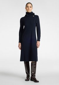 Abito lungo navy a maniche lunghe con ampio collo a rotondo, dettagli con bottoni frontali e silhouette dritta. Abbinato a stivali marroni alti fino al ginocchio con finitura lucida.