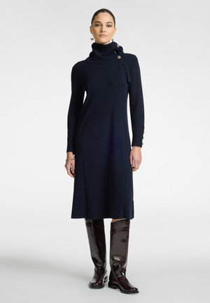 Navy lange mouw jurk met een brede rolkraag, frontknopen en een rechte pasvorm. Gecombineerd met hoogglanzende, bruine kniehoge laarzen.