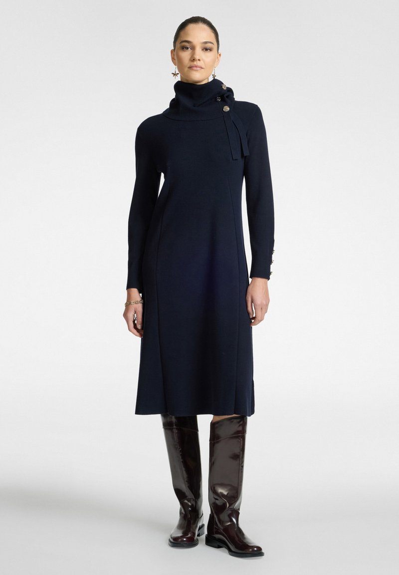 Abito lungo navy a maniche lunghe con ampio collo a rotondo, dettagli con bottoni frontali e silhouette dritta. Abbinato a stivali marroni alti fino al ginocchio con finitura lucida.