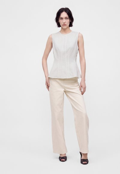 Femme aux cheveux courts et foncés, vêtue d'un haut blanc sans manches, d'un pantalon large couleur crème et de chaussures noires à bouts ouverts, debout devant un fond clair uni.