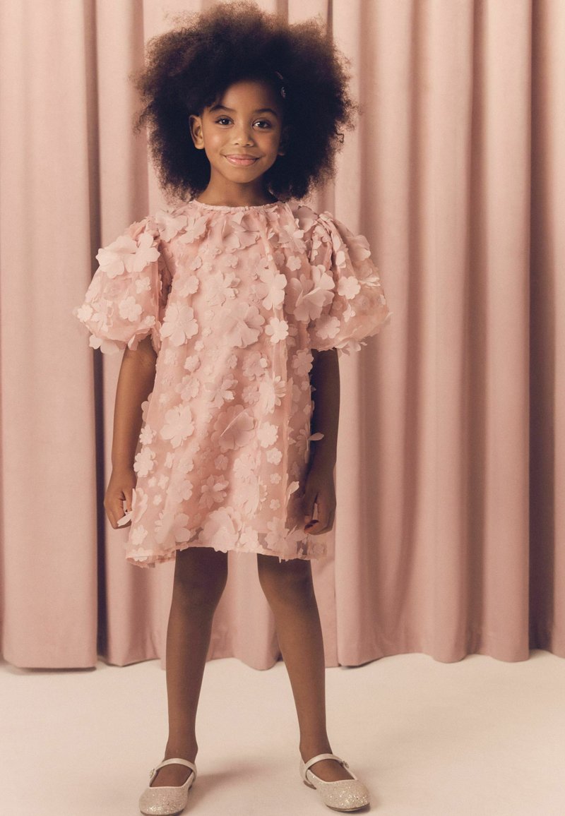 Junges Mädchen mit Afro-Frisur, das ein pinkes Kleid mit Blumenmuster und funkelnde Schuhe trägt, steht vor pinkfarbenen Vorhängen.